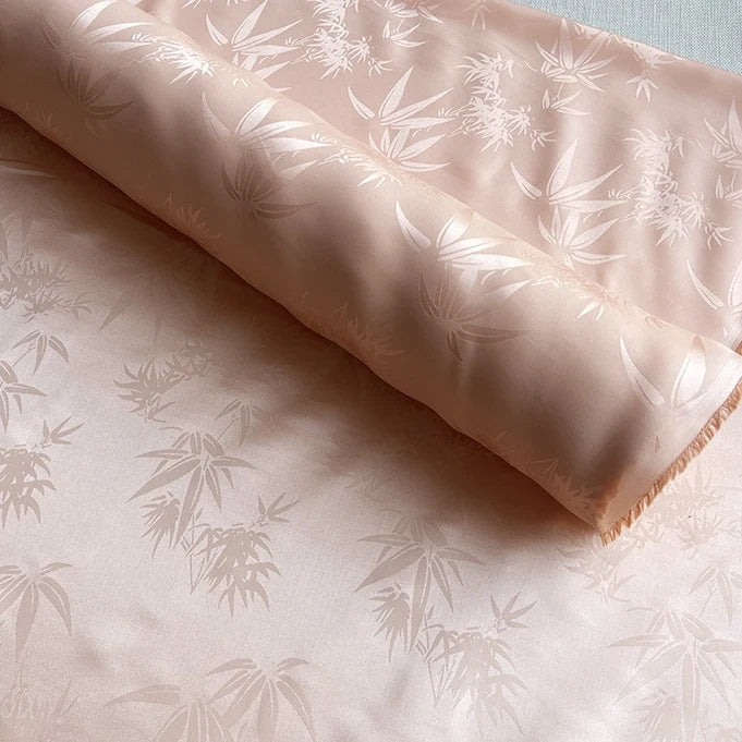 Un rouleau de Tissu soie de mûrier et bambou imprimé jacquard au mètre de Longan Craft France, un tissu de doublure doux et multicolore en soie-rayon avec un motif subtil de feuilles de bambou, est partiellement déroulé sur une surface plane.