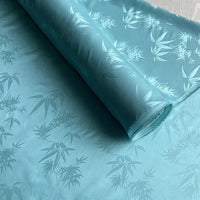 Un rouleau de tissu soie de mûrier et bambou imprimé jacquard de Longan Craft France est partiellement déroulé, révélant son motif de feuille de bambou doux et brillant, répété sur la doublure en soie et rayonne multicolore.