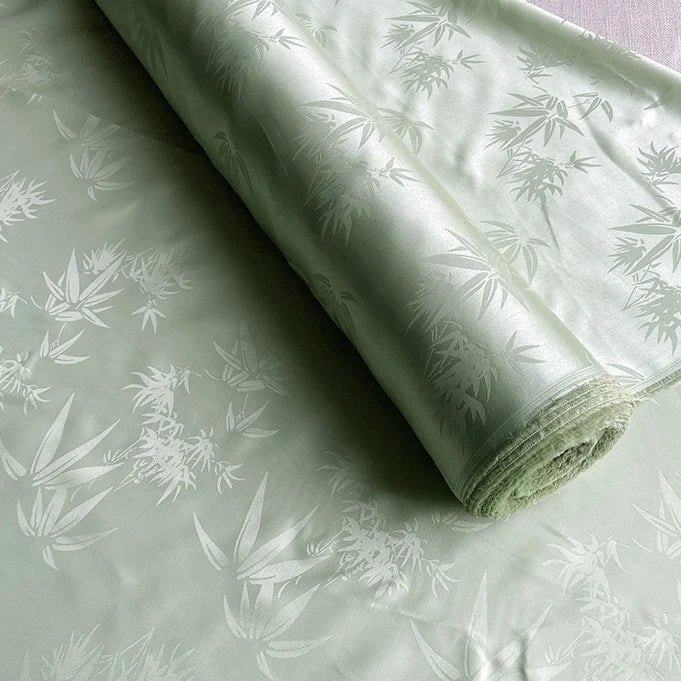 Un rouleau de Tissu soie de mûrier et bambou imprimé jacquard au mètre de Longan Craft France, présentant un subtil motif de feuilles de bambou et un léger éclat, est déroulé sur une surface plane. Le tissu soie-rayon est doux, coloré et luxueusement tissé.