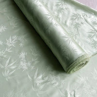 Un rouleau de Tissu soie de mûrier et bambou imprimé jacquard au mètre de Longan Craft France, présentant un subtil motif de feuilles de bambou et un léger éclat, est déroulé sur une surface plane. Le tissu soie-rayon est doux, coloré et luxueusement tissé.