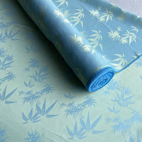 Un rouleau de Tissu soie de mûrier et bambou imprimé jacquard au mètre de Longan Craft France, bleu clair avec des motifs botaniques bleu pâle et jaune, est partiellement déroulé pour montrer ses délicats motifs de feuilles et de fleurs.