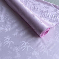 Un rouleau de tissu soie de mûrier et bambou imprimé jacquard au mètre de Longan Craft France, un mélange de soie-rayon rose pâle lisse et légèrement brillant avec un subtil motif de feuilles de bambou, est partiellement déroulé sur une surface plane.