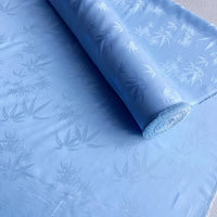 Un rouleau de tissu Longan Craft France soie de mûrier et bambou imprimé jacquard au mètre est partiellement déroulé, montrant son subtil motif de feuilles et ses reflets soyeux multicolores - idéal pour une doublure douce et confortable en soie et rayonne.