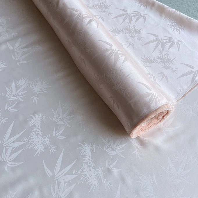 Un rouleau de tissu Longan Craft France soie de mûrier et bambou imprimé jacquard au mètre est partiellement déroulé, montrant son doux motif de feuilles de bambou multicolore en soie et rayonne avec des finitions mates et brillantes.