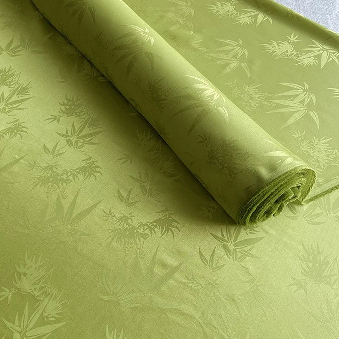 Un rouleau de tissu soie de mûrier et bambou imprimé jacquard au mètre de Longan Craft France, vert clair avec un subtil motif de feuilles de bambou, est partiellement déroulé ; ce tissu lisse et léger en soie-rayon a un éclat doux et est idéal comme doublure confortable.