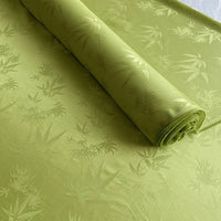Un rouleau de tissu soie de mûrier et bambou imprimé jacquard au mètre de Longan Craft France, vert clair avec un subtil motif de feuilles de bambou, est partiellement déroulé ; ce tissu lisse et léger en soie-rayon a un éclat doux et est idéal comme doublure confortable.