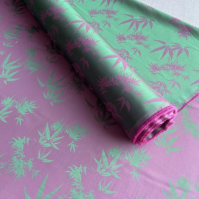 Un rouleau de tissu soie de mûrier et bambou imprimé jacquard au mètre de Longan Craft France, présentant un fond violet avec des feuilles de bambou vertes, est partiellement déroulé, montrant sa texture satinée brillante et son motif multicolore complexe.