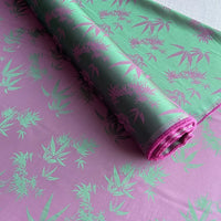 Un rouleau de tissu soie de mûrier et bambou imprimé jacquard au mètre de Longan Craft France, présentant un fond violet avec des feuilles de bambou vertes, est partiellement déroulé, montrant sa texture satinée brillante et son motif multicolore complexe.