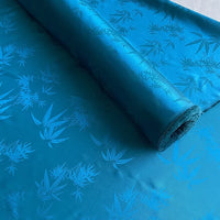 Un rouleau de Tissu soie de mûrier et bambou imprimé jacquard au mètre de Longan Craft France, un doux tissu de doublure en soie et rayonne multicolore avec un subtil motif de feuilles de bambou, est partiellement déroulé sur une surface plane.