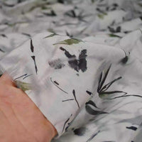 Une main tient un morceau de Tissu viscose imprimé bambou au mètre de Longan Craft France, une viscose légère style chinoiserie, idéale pour robes et confection, à motif floral noir et vert sur fond blanc semi-transparent.