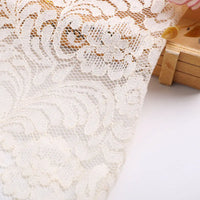 Gros plan sur la Dentelle florale beige au mètre de Longan Craft France, montrant son motif complexe de grandes fleurs et de feuilles. Cette dentelle ajourée est parfaite pour la confection de vêtements, drapée sur une surface en bois léger.