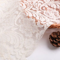Gros plan sur la Dentelle florale beige au mètre de Longan Craft France, avec ajouré grandes fleurs, drapée sur un panier en osier. Une pomme de pin brune est posée à côté du panier sur une surface blanche.