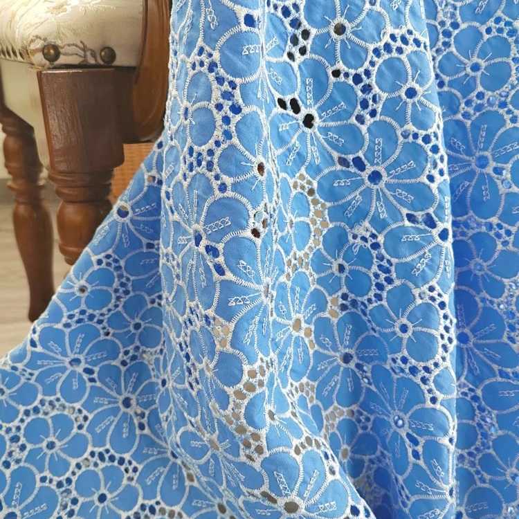Gros plan sur le Tissu brodé floral ajouré bleu au mètre de Longan Craft France, un tissu 100 % coton ajouré à fleurs, drapé sur une surface près d'une chaise en bois à assise claire.