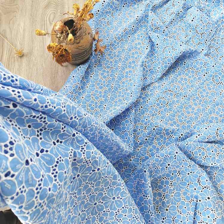Le Tissu brodé floral ajouré bleu au mètre de Longan Craft France, un tissu floral à œillets 100 % coton, est présenté drapé sur un plancher en bois à côté d'un vase en céramique contenant des fleurs jaunes séchées.