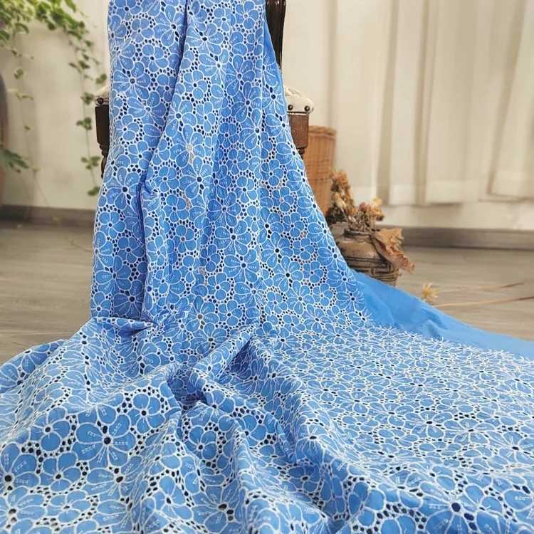 Un grand morceau de Tissu brodé floral ajouré bleu au mètre de Longan Craft France est posé sur le sol et sur une chaise, son motif de dentelle florale blanche se détachant sur des rideaux légers, de la verdure et un panier en osier dans ce cadre paisible.