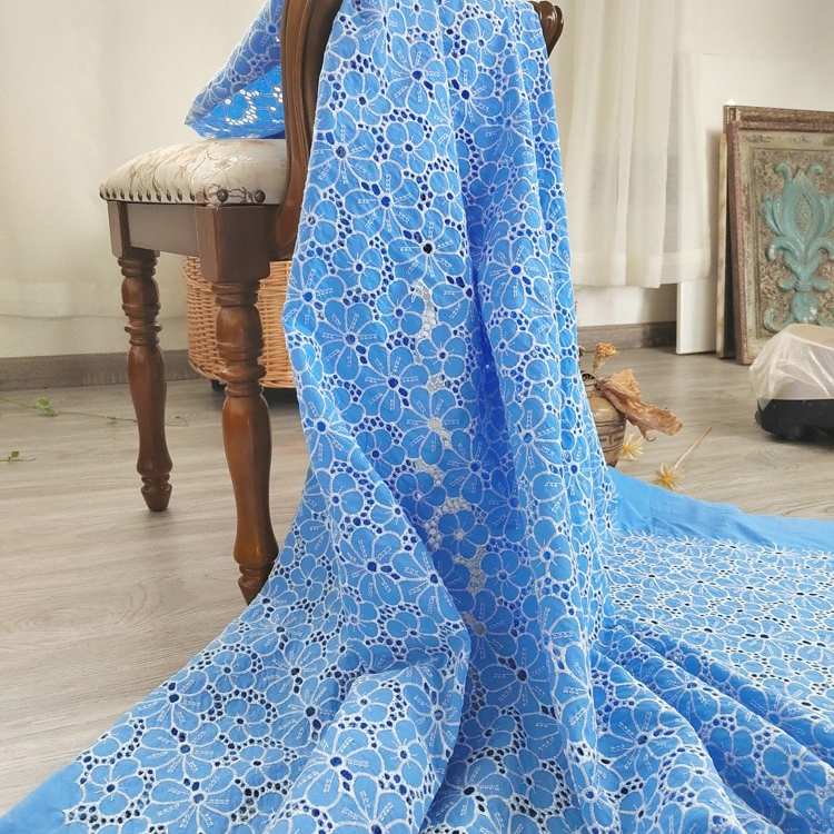 Un morceau de Tissu brodé floral ajouré bleu au mètre de Longan Craft France, 100 % coton avec découpe florale, est drapé sur une chaise en bois avec coussin, posée sur un plancher en bois clair dans une pièce aux rideaux et à la décoration blancs.