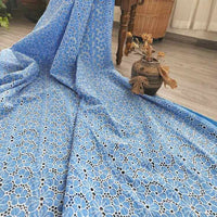 Un grand morceau de tissu brodé floral ajouré bleu au mètre de Longan Craft France est étalé sur un sol en bois, avec un vase en céramique contenant des feuilles séchées à proximité, ainsi qu'un panier en osier et un rideau à l'arrière-plan.