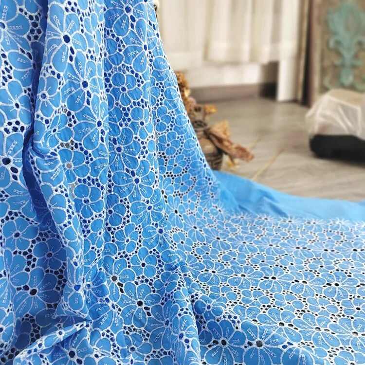 Une longueur de "Tissu brodé floral ajouré bleu au mètre" de Longan Craft France, un tissu à œillets floral 100 % coton, est drapée sur une surface avec une partie d'une pièce et un rideau à l'arrière-plan.