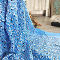 Une longueur de "Tissu brodé floral ajouré bleu au mètre" de Longan Craft France, un tissu à œillets floral 100 % coton, est drapée sur une surface avec une partie d'une pièce et un rideau à l'arrière-plan.