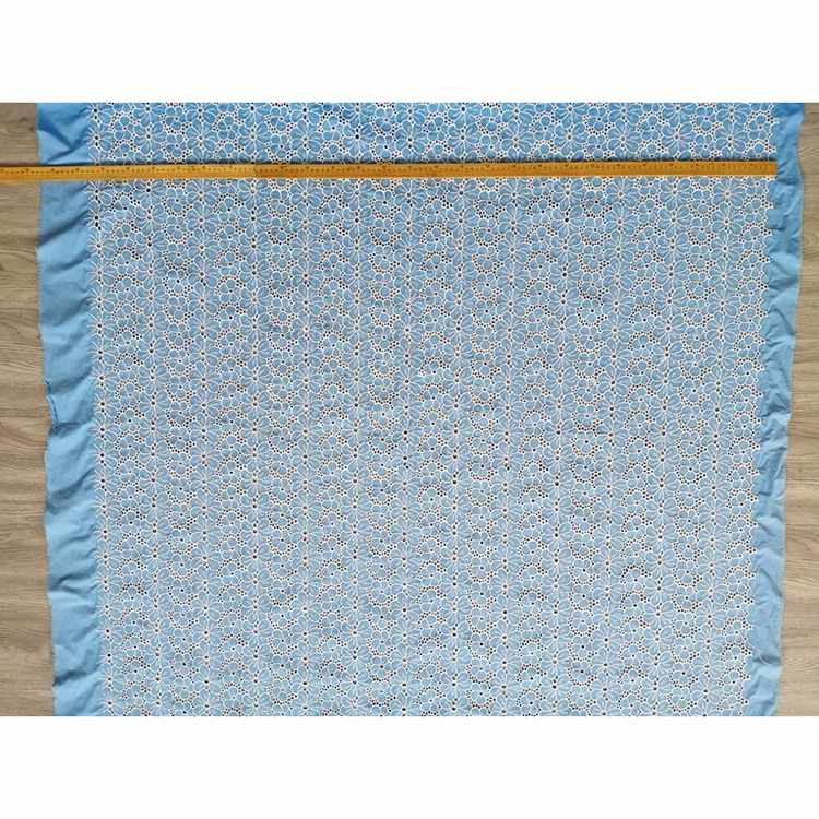 Un tissu brodé floral bleu en coton de Longan Craft France est posé à plat sur du bois, avec un mètre le long du bord supérieur. Ce "tissu brodé floral ajouré bleu au mètre" est 100 % coton et présente un motif floral ajouré.