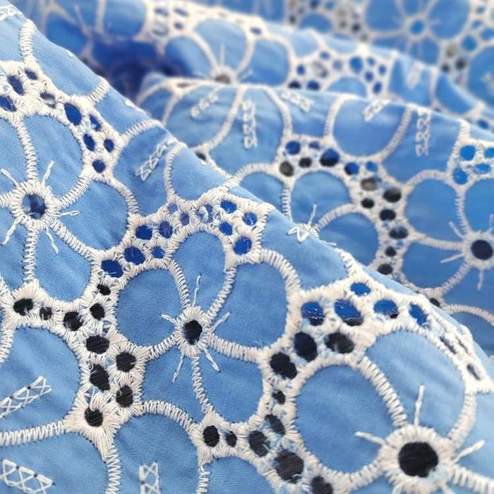 Gros plan sur le Tissu brodé floral ajouré bleu au mètre de Longan Craft France, un tissu 100% coton ajouré à base de bleu clair et de broderies florales blanches et bleu foncé, mettant en valeur les coutures détaillées et les motifs circulaires texturés.