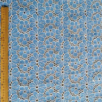 Le Tissu brodé floral ajouré bleu au mètre de Longan Craft France est un tissu 100 % coton, qui présente un motif de fleurs bleu clair répétées, soulignées en blanc et centrées en noir. Une règle en bois sur le bord indique les mesures en centimètres.