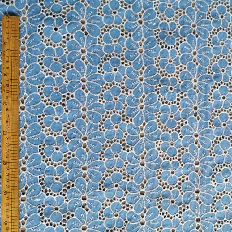 Le Tissu brodé floral ajouré bleu au mètre de Longan Craft France est un tissu 100 % coton, qui présente un motif de fleurs bleu clair répétées, soulignées en blanc et centrées en noir. Une règle en bois sur le bord indique les mesures en centimètres.