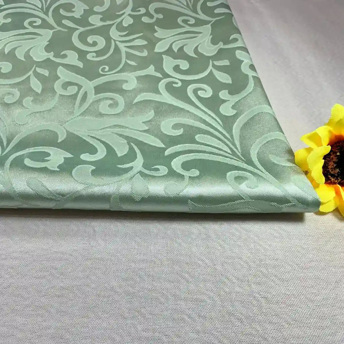 Un morceau plié du brocart au mètre Tissu damas de Longan Craft France, avec un subtil motif jacquard de fleurs et de feuilles, repose sur une surface claire, à côté d'une fleur artificielle jaune sur la droite.