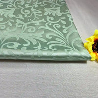 Un morceau plié du brocart au mètre Tissu damas de Longan Craft France, avec un subtil motif jacquard de fleurs et de feuilles, repose sur une surface claire, à côté d'une fleur artificielle jaune sur la droite.