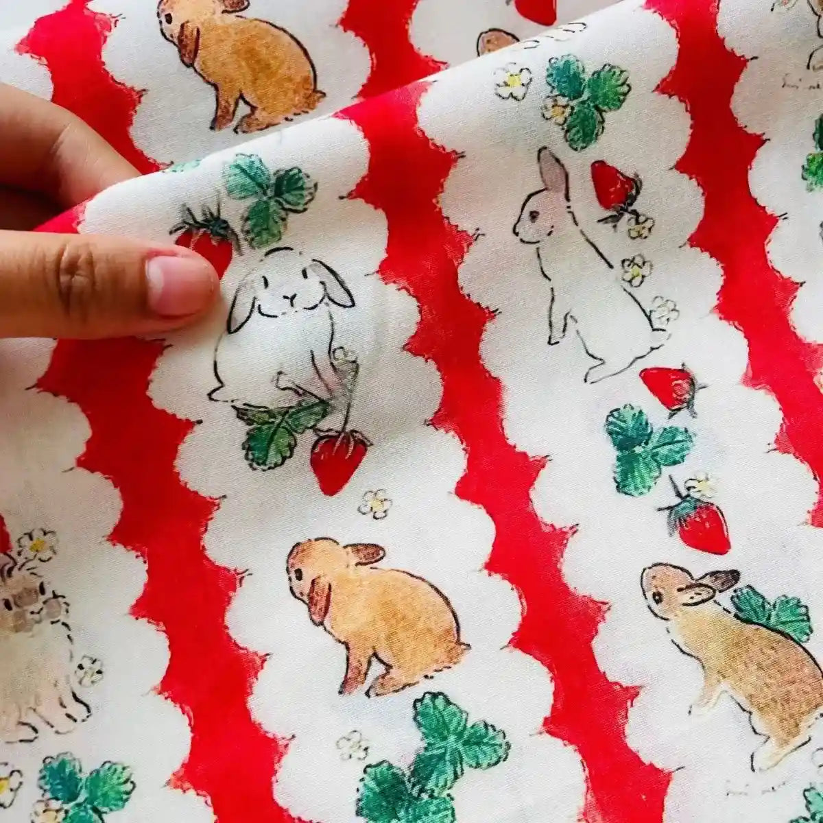 Une main tient le Tissu de Pâques motif lapin et carotte en coton au mètre de Longan Craft France, 100% coton doux et léger avec des lapins espiègles - parfait pour les projets de bricolage saisonniers.