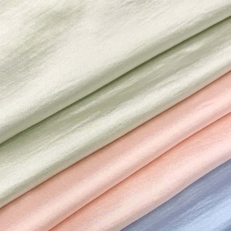 Gros plan sur quatre pièces pliées du tissu en soie charmeuse de mûrier de Longan Craft France, un mélange de rayonne et de polyester lisse et légèrement brillant dans des tons pastel blanc cassé, vert clair, pêche et bleu, idéal pour créer des robes fluides.
