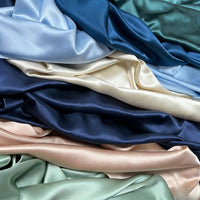 Plusieurs pièces de Tissu de soie charmeuse au mètre de Longan Craft France en bleu, vert, crème et pêche sont superposées, mettant en valeur la texture lisse et brillante de cette luxueuse soie de mûrier pour robes de mariée.
