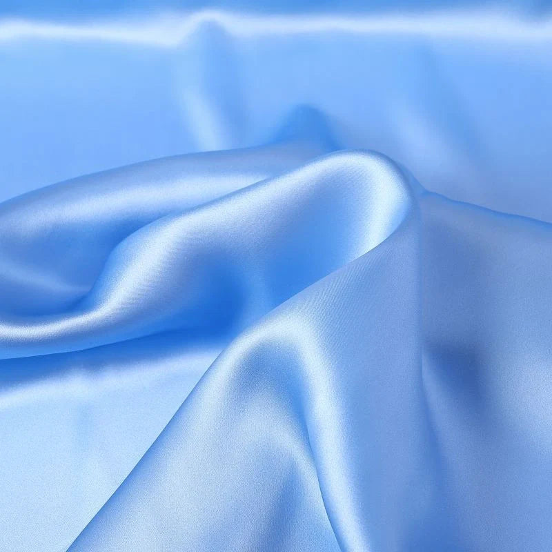 Un gros plan de Tissu de soie charmeuse au mètre, bleu clair et lisse, de Longan Craft France, montre des plis doux et des reflets délicats, mettant en valeur la luxueuse soie de mûrier, idéale pour les robes de mariée.