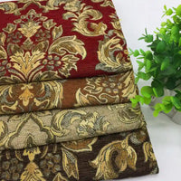 Quatre pièces pliées de tissu d'ameublement jacquard damassé de Longan Craft France, avec des motifs floraux rouges, bruns, beiges et marron foncé, sont empilées sur une surface blanche à côté d'une petite plante verte en pot.