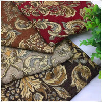 Trois pièces pliées en tissu damassé jacquard à motifs floraux rouge, marron, beige et or de Longan Craft France sont empilées à côté d'une petite plante verte en pot.