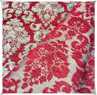 Gros plan sur le tissu damassé chenille au mètre de Longan Craft France, un jacquard d'ameublement aux motifs floraux ornés de rouge et d'or. Un coin est plié pour révéler le motif complexe au verso.