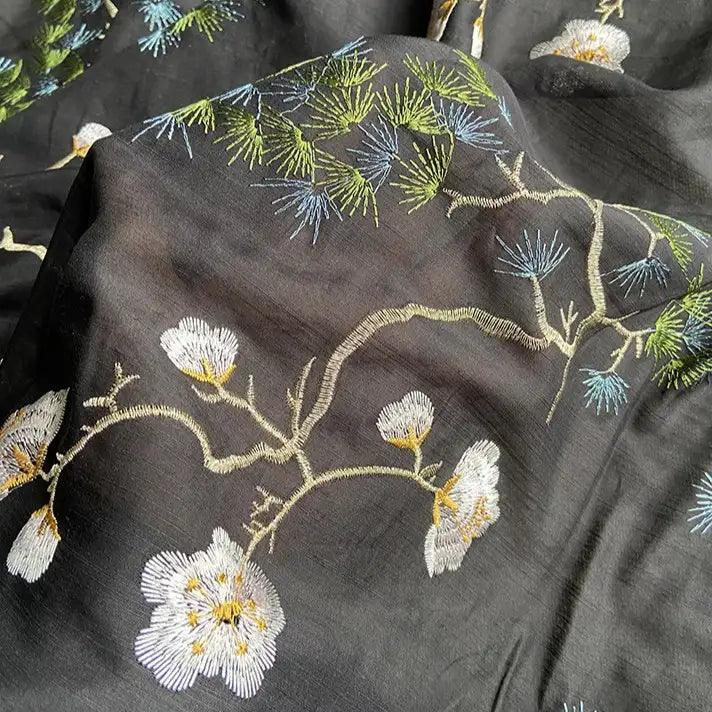 Le tissu coton et soie brodé de style chinoiserie de Longan Craft France est un tissu noir brodé de fleurs blanches, de centres jaunes, de feuilles vertes et d'aiguilles de pin bleues - un tissu floral en soie de mûrier de première qualité, parfait pour les chemisiers.