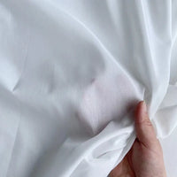 Une main tient un morceau de Longan Craft France soie de coton multicolore au mètre - un tissu de doublure en soie de mûrier léger et respirant - en le tirant légèrement pour révéler sa qualité fine et semi-transparente ainsi qu'un léger contour de la main.