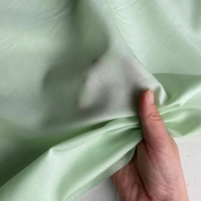 Une main pince un morceau de "Soie de coton multicolore au mètre | Soie de mûrier légère" vert clair, semi-transparent, de Longan Craft France, les ombres des doigts étant visibles à travers le tissu délicat et respirant de la doublure en soie.