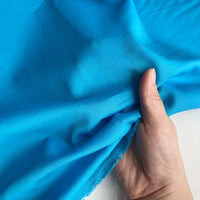 Une main soulève une Soie de coton multicolore au mètre de Longan Craft France, révélant la texture légère et translucide de cette soie de mûrier utilisée comme tissu de doublure en soie respirant sur un fond clair.