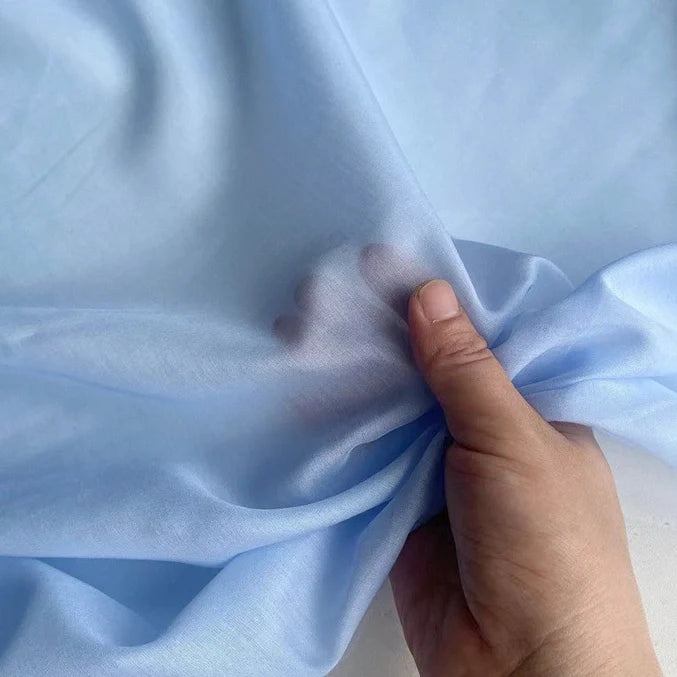 Une main rassemble un morceau de tissu bleu clair, transparent, soulignant sa texture aérienne - idéal pour mettre en valeur la qualité délicate de Soie de coton multicolore au mètre de Longan Craft France, un tissu de doublure en soie de mûrier léger et respirant.