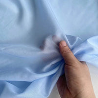Une main rassemble un morceau de tissu bleu clair, transparent, soulignant sa texture aérienne - idéal pour mettre en valeur la qualité délicate de Soie de coton multicolore au mètre de Longan Craft France, un tissu de doublure en soie de mûrier léger et respirant.