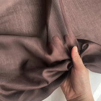 Une main tient et froisse délicatement le Soie de coton multicolore au mètre de Longan Craft France, un tissu de doublure en soie léger et semi-transparent. Le fond uni met en valeur la texture délicate et respirante de la soie de mûrier.