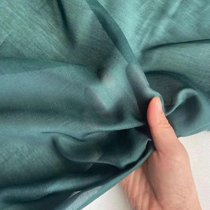 Une main tient et rassemble délicatement Soie de coton multicolore au mètre de Longan Craft France - un tissu de doublure en soie de mûrier léger et respirant - montrant sa texture fine et sa translucidité sur un fond clair.