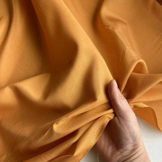 Une main froisse délicatement un morceau de Soie de coton multicolore au mètre de Longan Craft France, mettant en valeur ce tissu de doublure en soie de mûrier, léger et respirant, en orange clair sur un fond neutre.