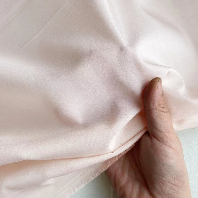 Une main tient et pince délicatement un morceau de Soie de coton multicolore au mètre de Longan Craft France, révélant la texture douce et l'éclat subtil de la doublure en soie de mûrier, légère et respirante, à la lumière naturelle.