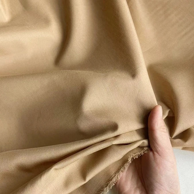 Une main tient et froisse délicatement le tissu Soie de coton multicolore au mètre de Longan Craft France - un tissu de soie mélangée de couleur mûre claire - révélant sa texture douce et respirante avec des plis naturels et un bord effiloché.