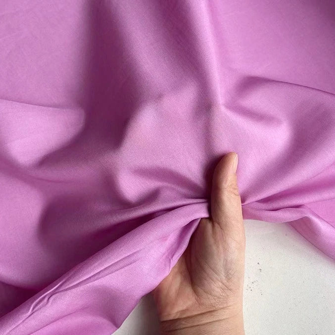 Une main pince délicatement un morceau de Soie de coton multicolore au mètre de Longan Craft France, un tissu de doublure en soie mélangée de couleur mûre, montrant sa texture lisse sur un fond uni.