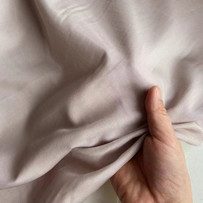 Une main rassemble un morceau de Soie de coton multicolore au mètre de Longan Craft France, un mélange léger de soie et de coton de couleur mûre à la texture lisse et au lustre subtil, parfait comme tissu de doublure en soie respirante.