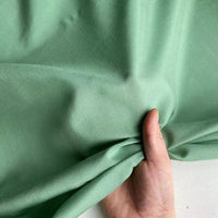 Une main rassemble un morceau de Soie de coton multicolore au mètre de Longan Craft France, un tissu de doublure en soie de mûrier clair qui montre sa texture lisse et légèrement froissée. L'arrière-plan est légèrement flou et neutre.